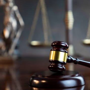 A gavel rests on a wooden table. Out of focus in the background are the scales of justice.