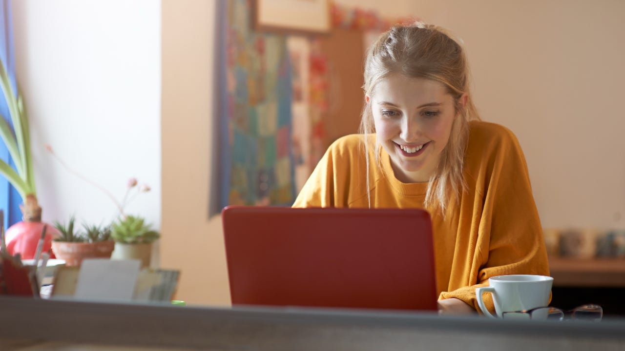 Woman in an orange shirt sitting at table while smiling at her laptop.