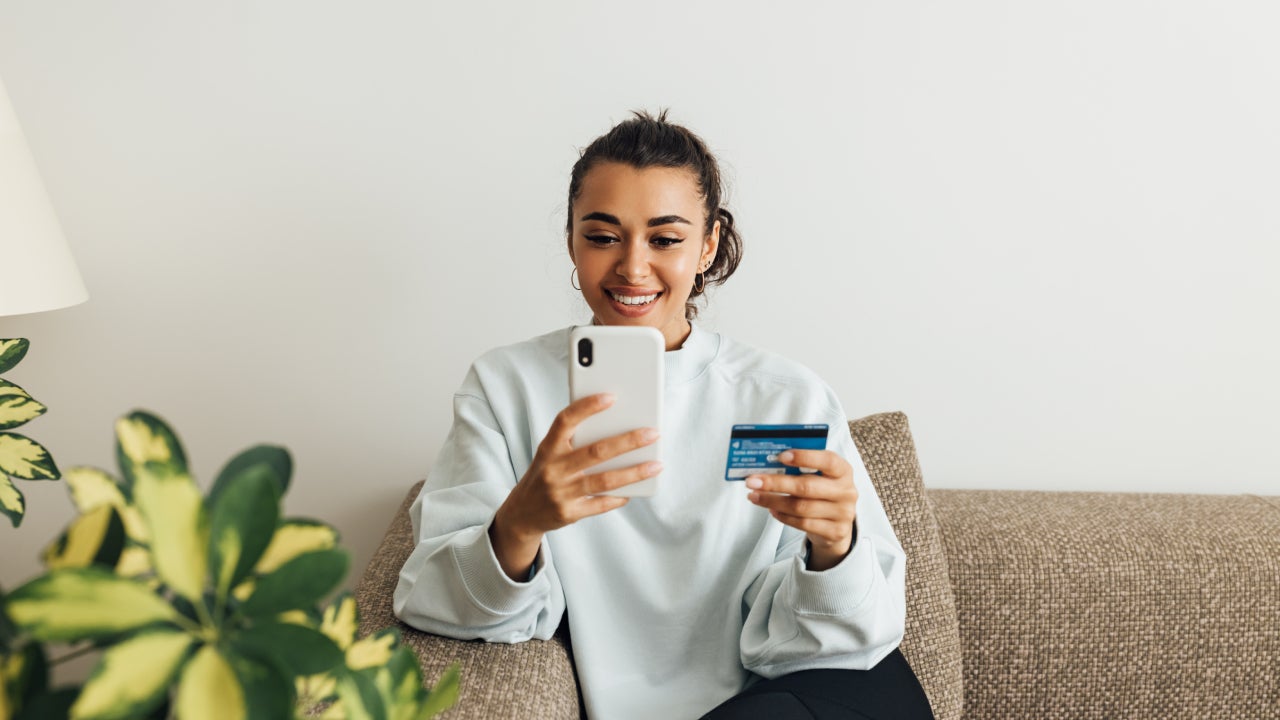 Smiling Woman Holding Credit Card While Using Smart Phone At Home - stock photo