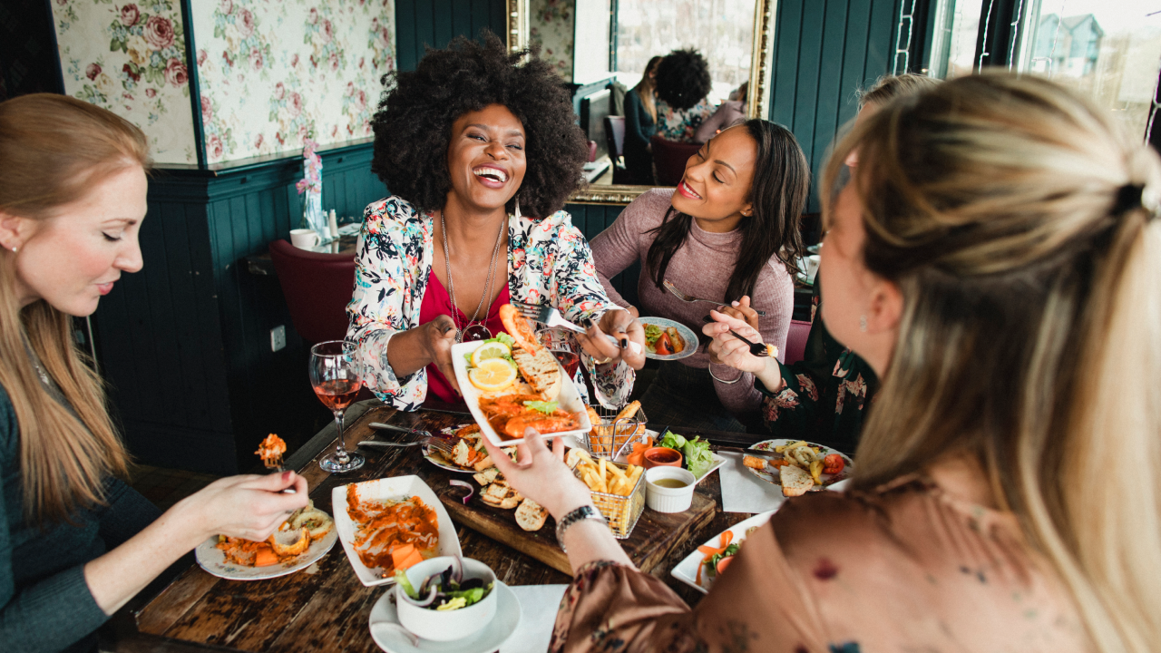 A group of women are enjoying a meal in a restaurant with rose wine. They have a king prawns sharing platter , one woman can be seen passing her friend a plate of food. Mixed ethnic group