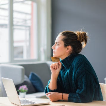 Profile of pensive woman with laptop and credit card sitting at desk at home looking out of window