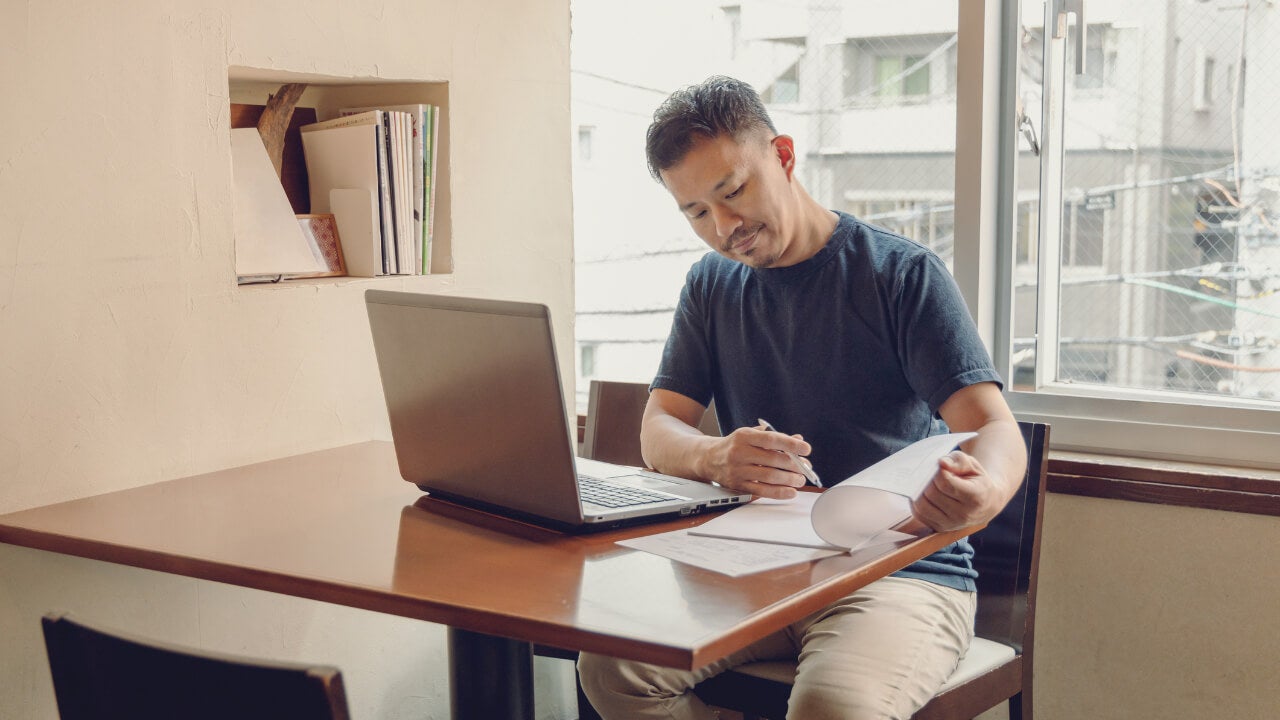 A Japanese man sits at a table with a laptop, looking at documents.