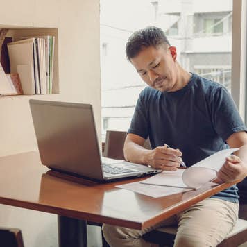 A Japanese man sits at a table with a laptop, looking at documents.