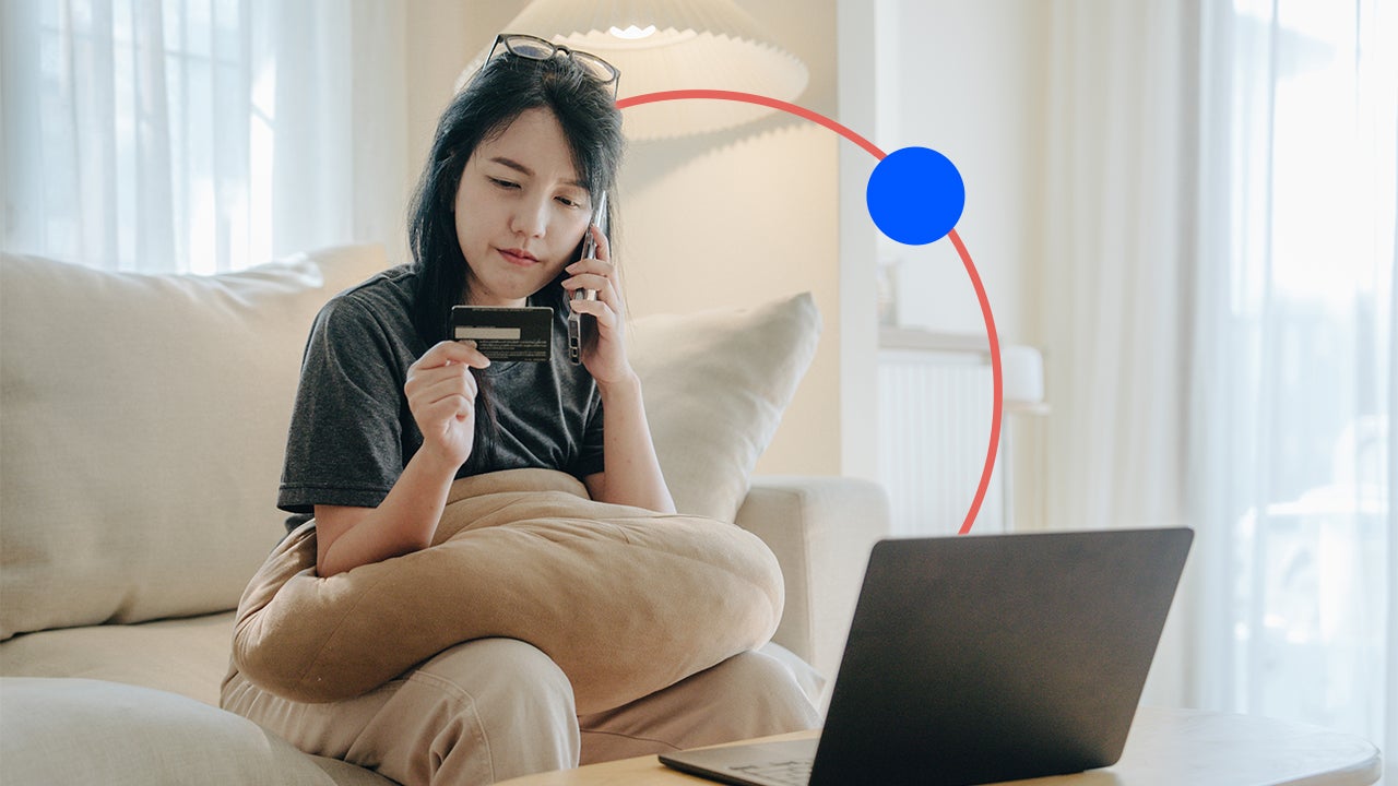 design image of a woman on the phone holding a credit card in one hand and sitting in front of a laptop on a couch