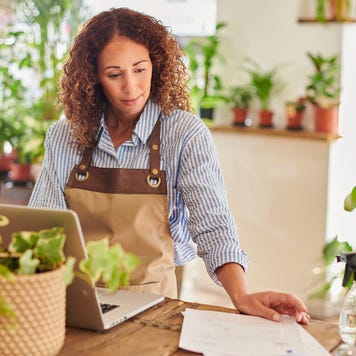 A small business owner stands in her flower shop, looking over her business accounts on her laptop.