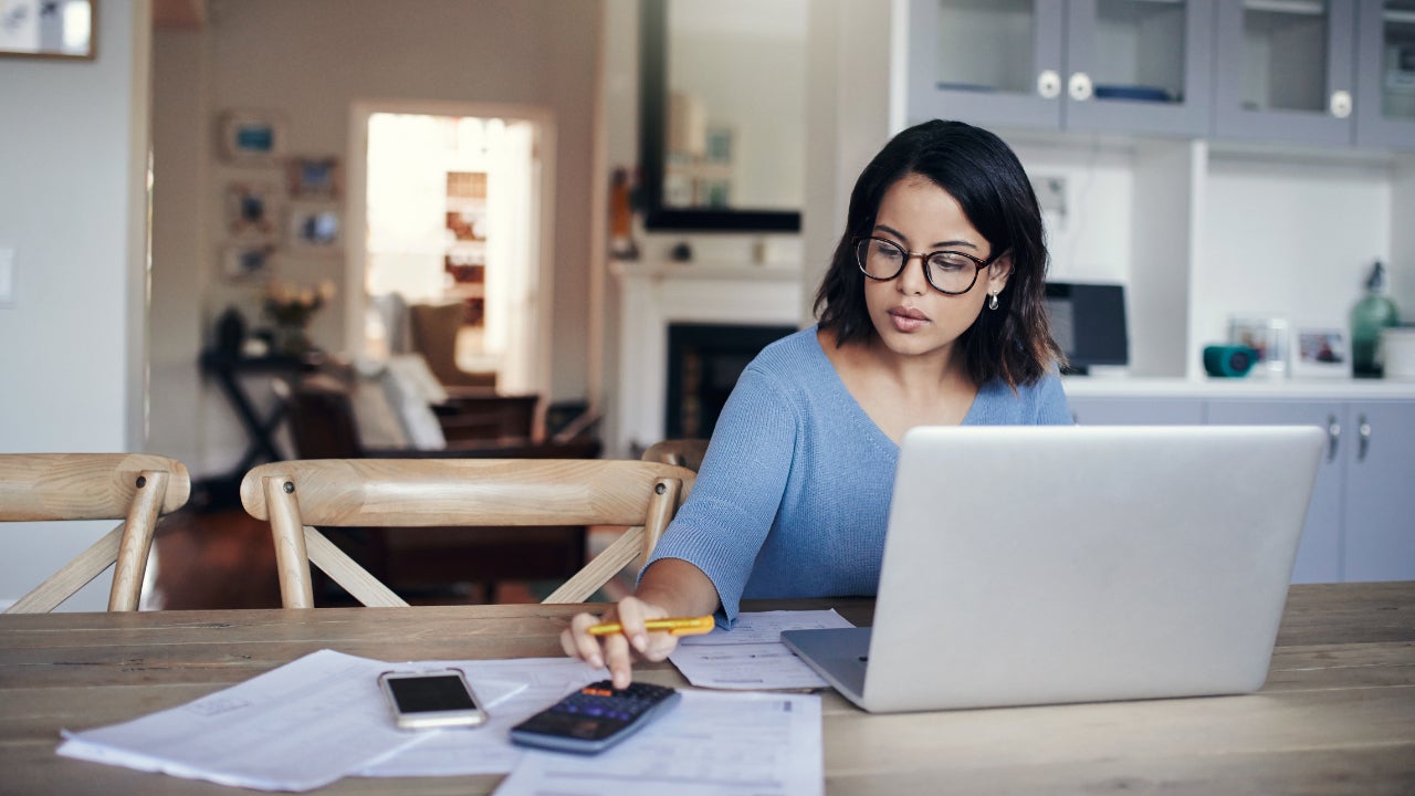 A woman sits at her kitchen table with her laptop, spreadsheets and calculator
