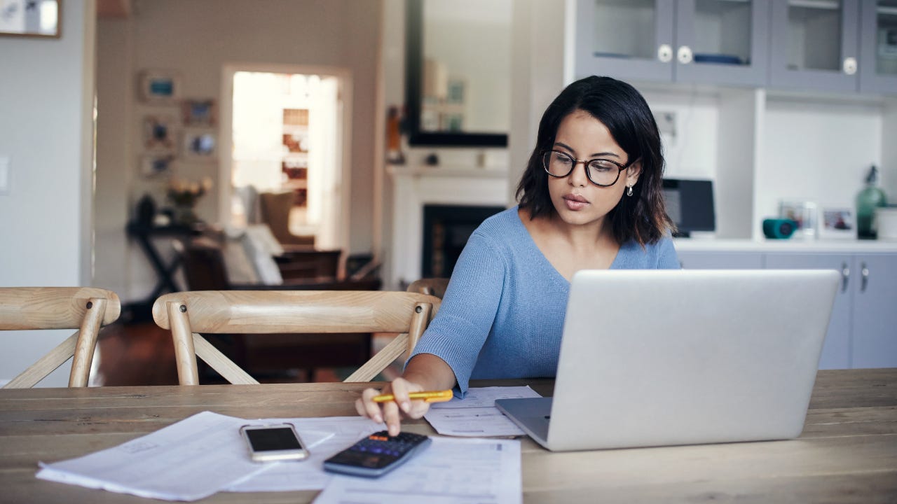 A woman sits at her kitchen table with her laptop, spreadsheets and calculator