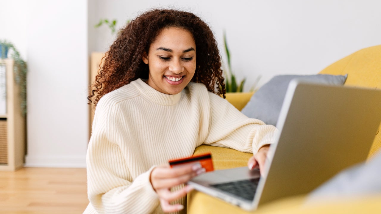 Smiling young ethnic woman holding credit card and using laptop computer at home. Online shopping concept
