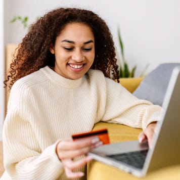 Smiling young ethnic woman holding credit card and using laptop computer at home. Online shopping concept