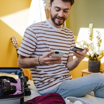 Young smiling man in a striped t-shirt using mobile phone and holding credit card at bedroom. Getting ready for a vacation.