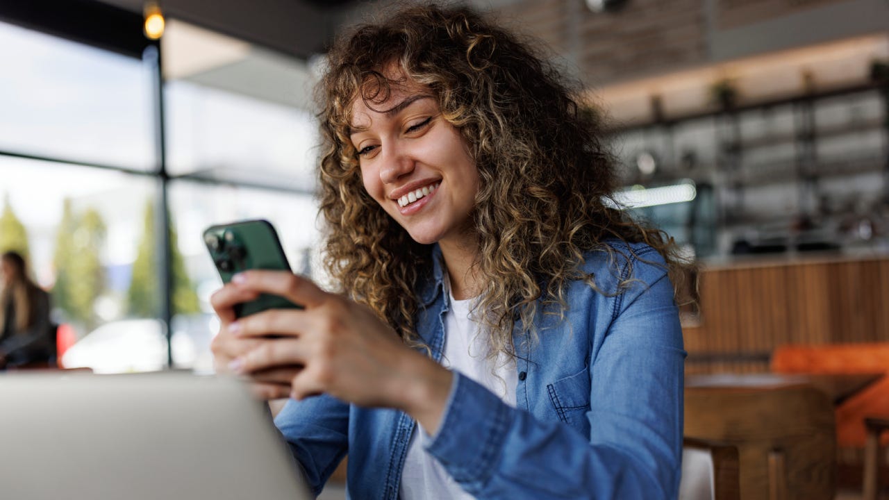 Young smiling woman using mobile phone while working on a laptop at a cafe