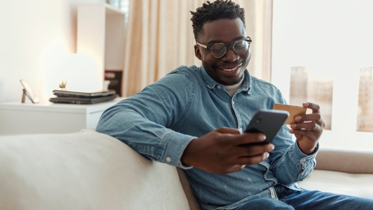 Shot of a young man holding his credit card while using a smartphone at home. Cheerful young man doing online shopping while using his cellphone and credit card inside of an apartment during the day.