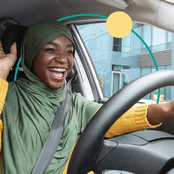A Black woman wearing a green hijab and yellow sweater smiles happily while inside of a car.