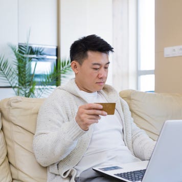 young asian man shopping online in internet store using laptop computer and credit card. male enters data of paying for services or tickets in application sitting on couch at home living room