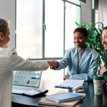 A lender shakes the hand of a smiling business owner who sits next to her partner in the lender’s office.