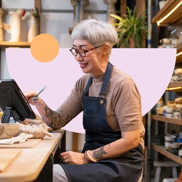 Older woman with tattoos works on a tablet in a ceramic studio.