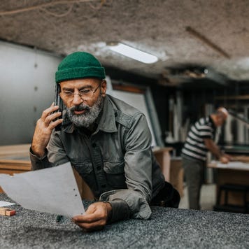Man with beard looks over paperwork while talking on the phone in a workshop.