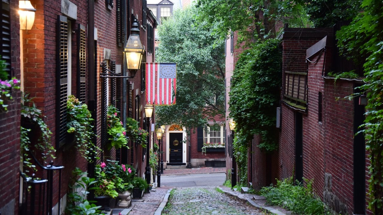 The narrow cobblestoned Acorn Street with Federal-style red row houses, with brick sidewalks and lit by gas lanterns at dawn, with an American flag in the historical neighbourhood of Beacon Hill