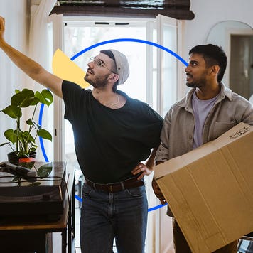 Two men standing together inside a house. One is holding a box.
