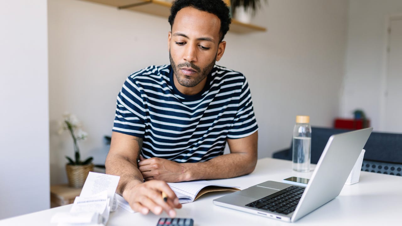 Young Black man using a calculator with a laptop open.