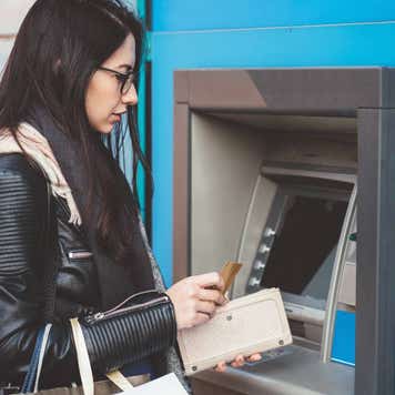 A person in casual attire stands at an ATM machine, inserting a card and interacting with the keypad to carry out a transaction.