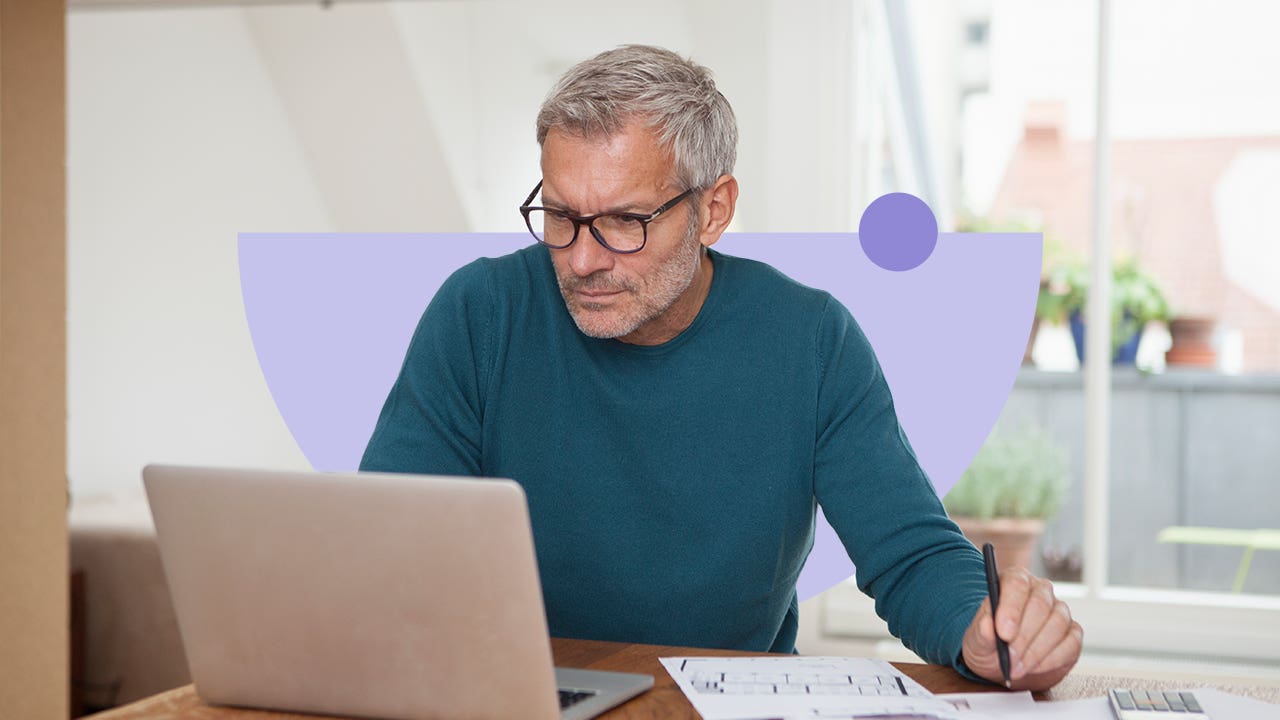 Older white man uses a laptop while writing on a piece of paper.