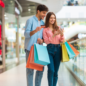 Shopping Application. Happy Husband And Wife Using Smartphone And Credit Card Holding Colorful Shopper Bags And Using Mobile App Buying Clothes Online In Modern Mall Indoor. Ecommerce Concept