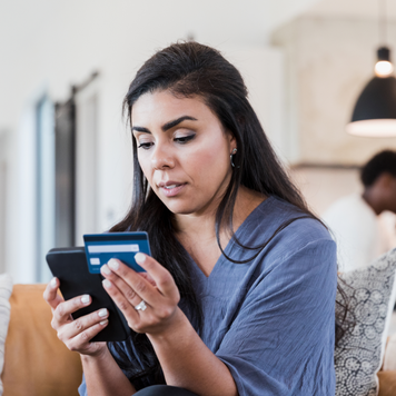 A serious mid adult woman enters her credit card number while shopping online. Her husband is working in the background.
