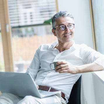 Thoughtful businessman holding coffee cup and laptop looking out through window at office