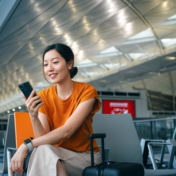 Young Asian woman with suitcase using smartphone while waiting for her flight at airport terminal. Asian businesswoman on business travel. Lifestyle and technology. Travel and vacation concept