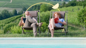 An older white couple sits together outside while reading books.