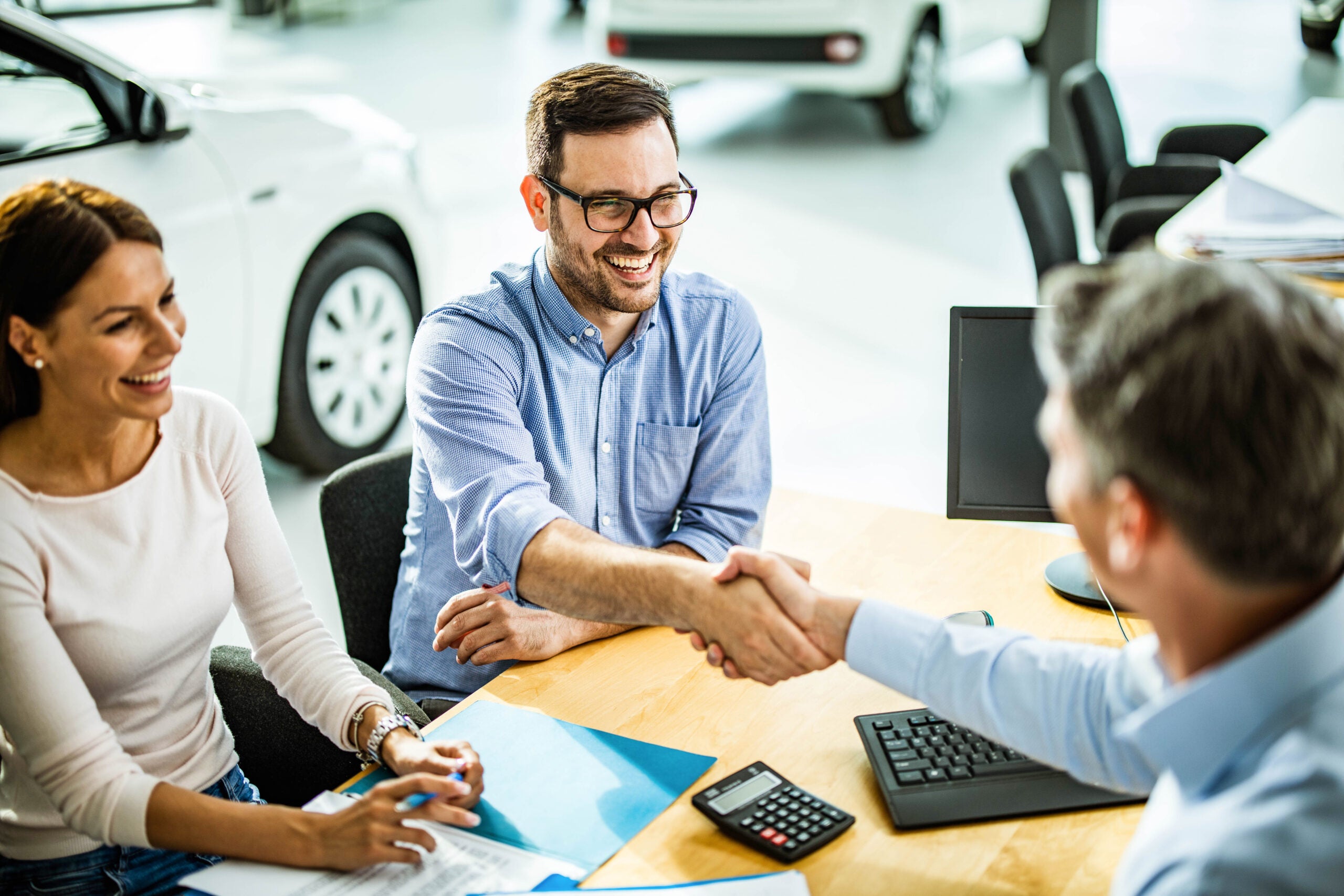 man and woman at a car dealership shaking hands with a salesperson after buying a car