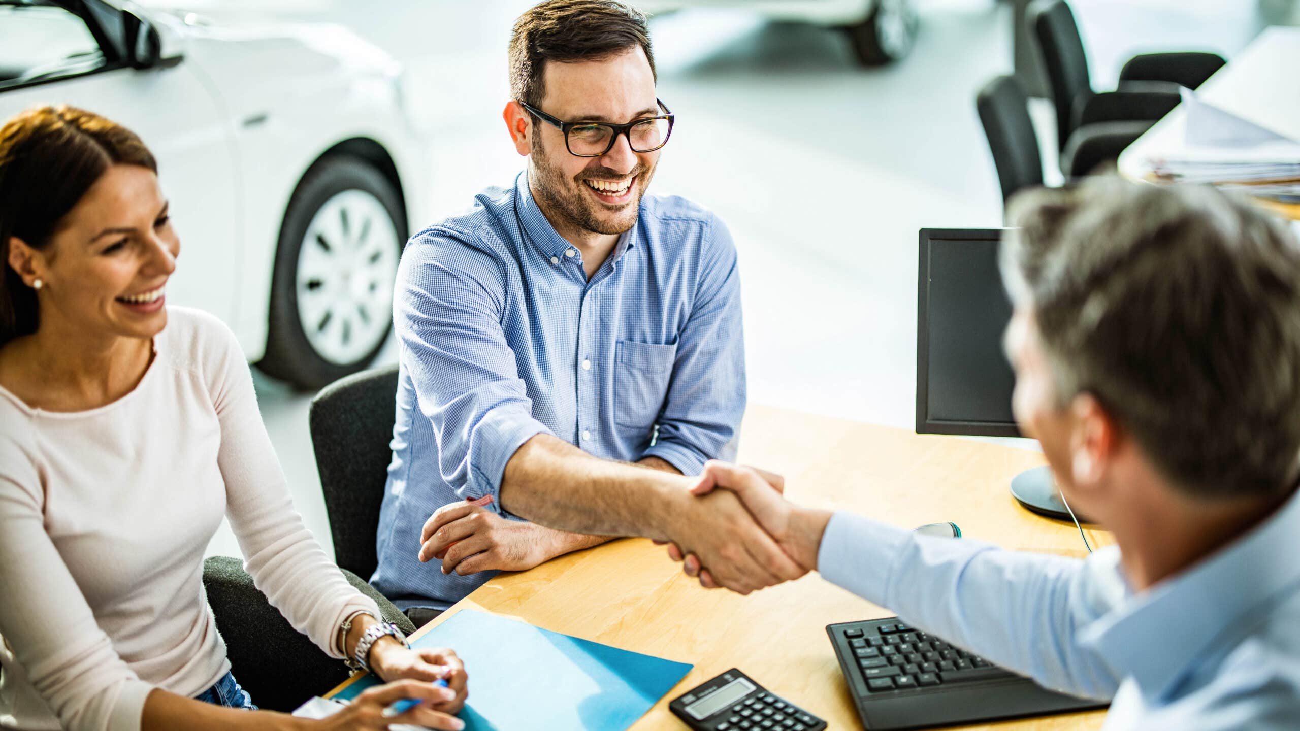 man and woman at a car dealership shaking hands with a salesperson after buying a car