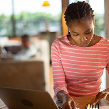Young Black woman in an orange shirt uses a calculator.