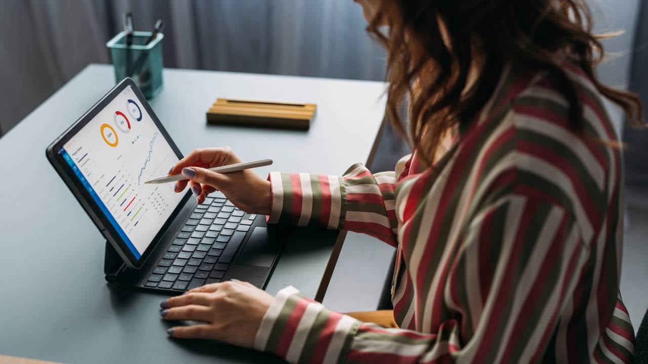 Focused young female analyzing financial statistics on digital tablet, working from a modern home office setup.