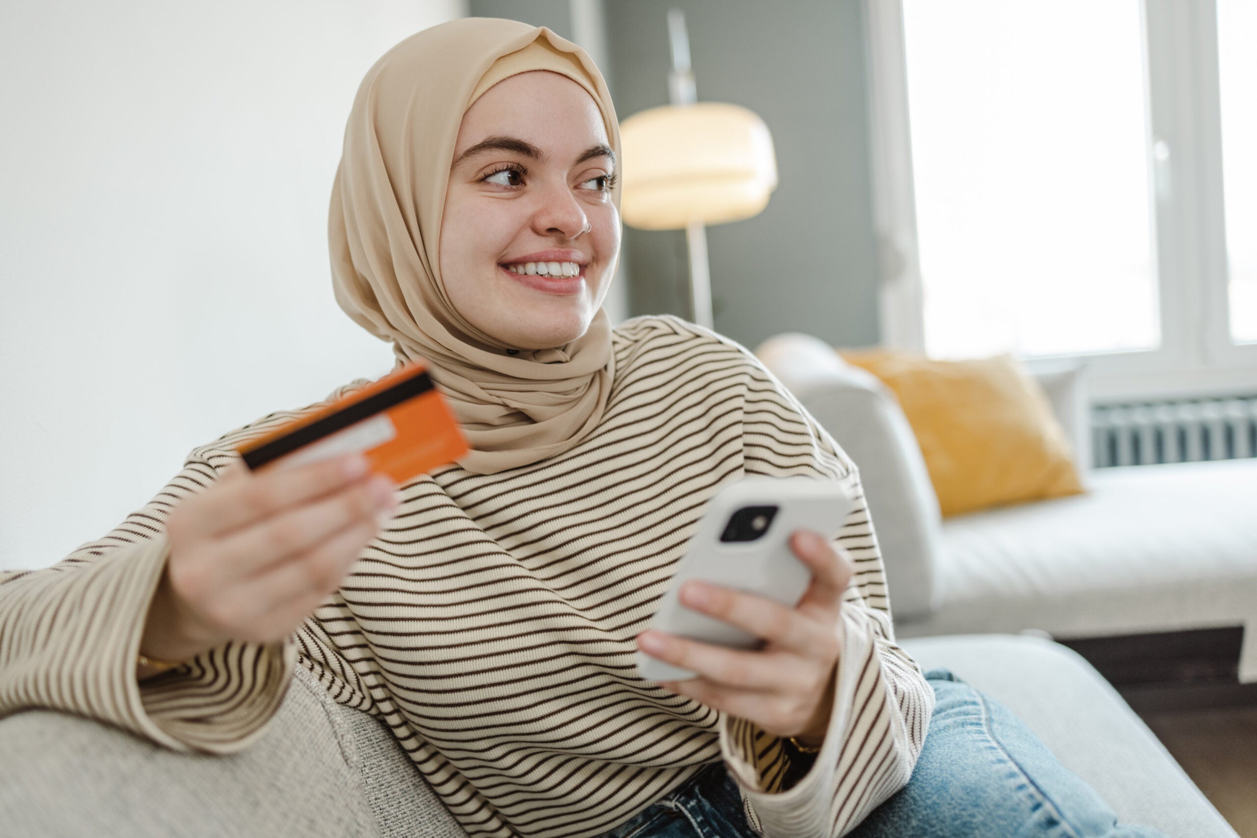 Photo of a smiling young woman holding a credit card and using a smart phone for online shopping