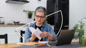 An older woman with black glasses on reviews papers at her table. There is an illustrated semicircle behind her.