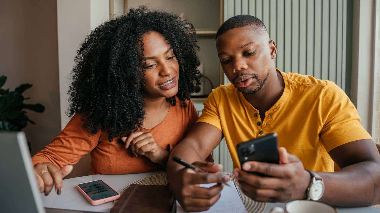 A young African-American man and woman sit at a table working on their finances with a laptop, phone and notebooks.