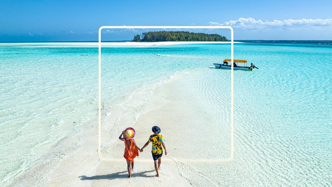 Image of two people in wide brim hats and summer clothes walking hand-in-hand down a narrow strip of white sand beach towards a small motor boat in the distance. Blue water laps in small waves on either side of the strip of beach.