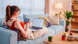 A young woman sits on her couch and trades stocks on her iPhone and computer.