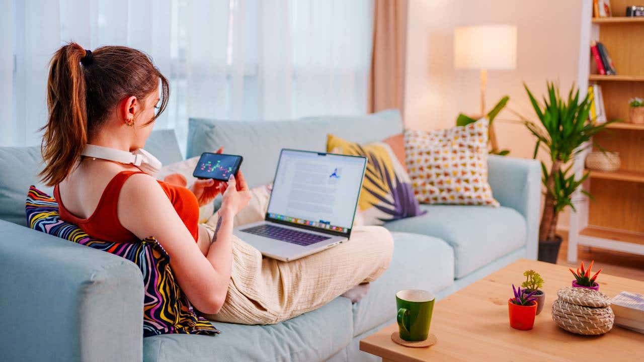 A young woman sits on her couch and trades stocks on her iPhone and computer.