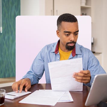 A Black man sits at a table using a tablet and doing calculations.