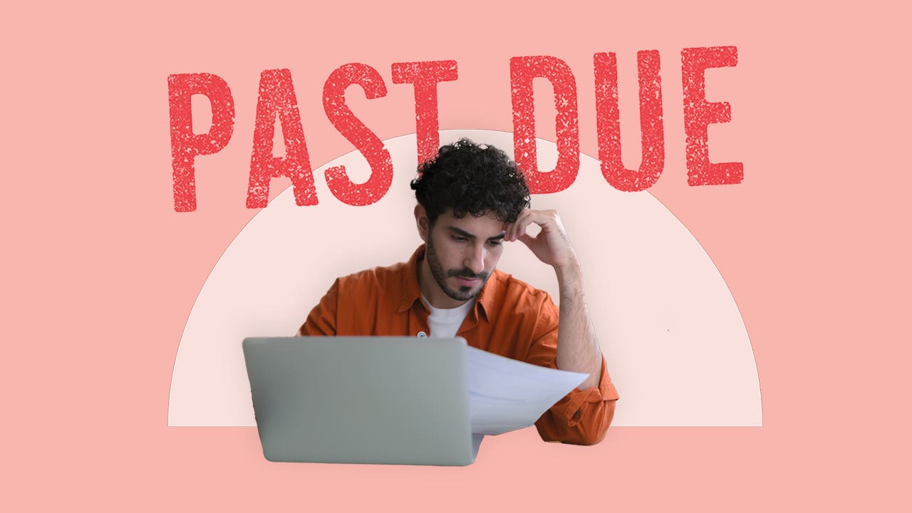 A man sitting in front of an open laptop computer looking down at some papers in his right hand. His left elbow is on the table and his head is leaning on his left hand.