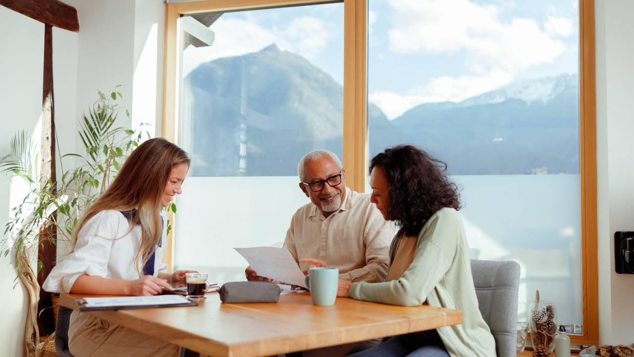 A financial advisor is seated with a senior couple in a bright room with a stunning mountain view. They are engaged in conversation, with documents laid out on the table, pointing to a discussion about pension insurance. The advisor is presenting information, while the couple listens intently, considering the options for their retirement planning.