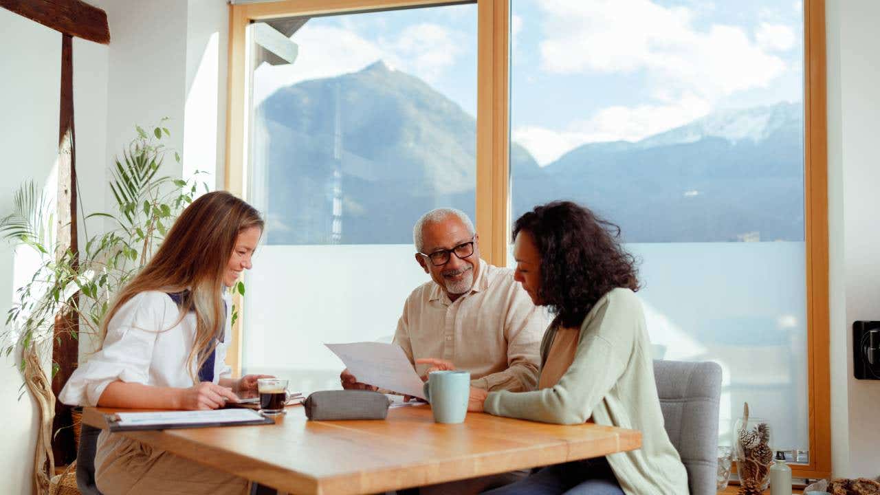 A financial advisor is seated with a senior couple in a bright room with a stunning mountain view. They are engaged in conversation, with documents laid out on the table, pointing to a discussion about pension insurance. The advisor is presenting information, while the couple listens intently, considering the options for their retirement planning.