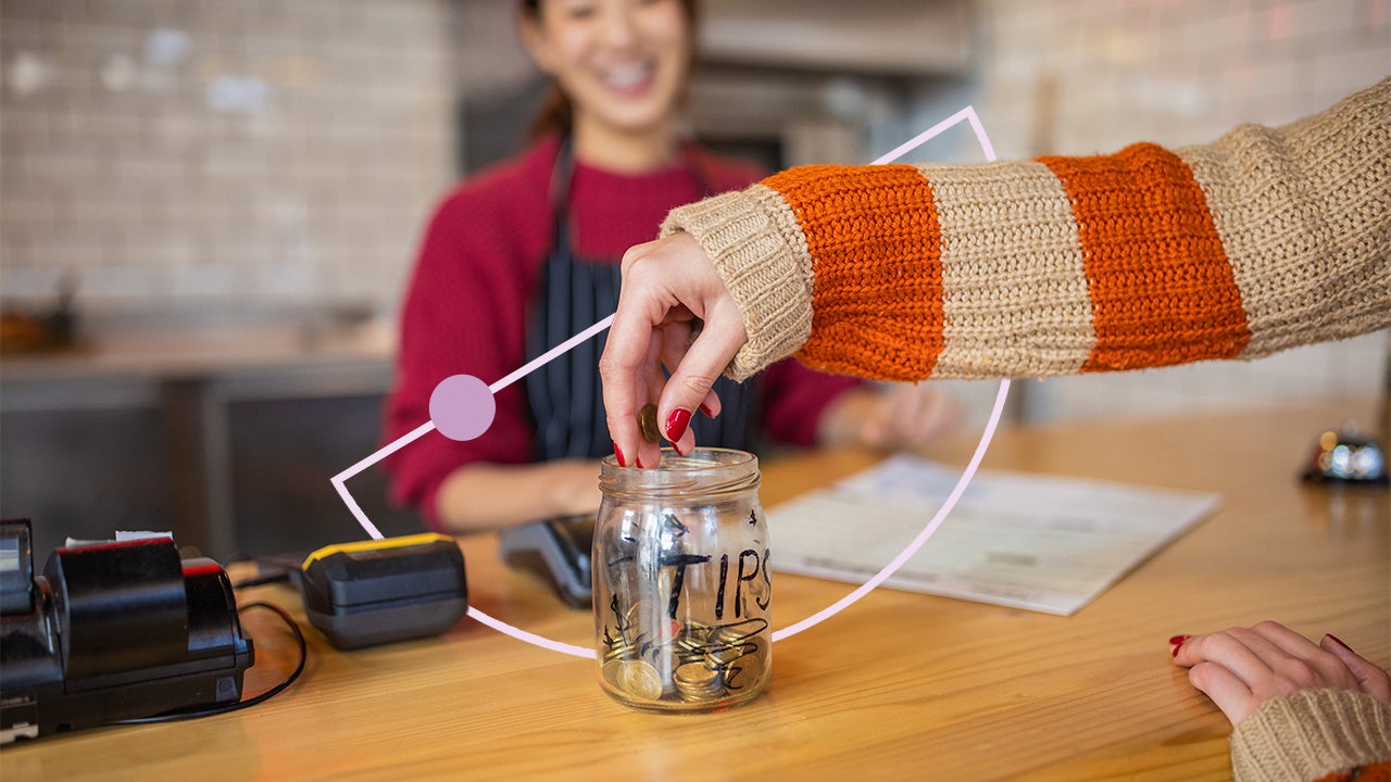 Image of a tip jar, filled with money, sitting on a counter. There is a hand adding more money to it while a smiling worker in an apron looks on.