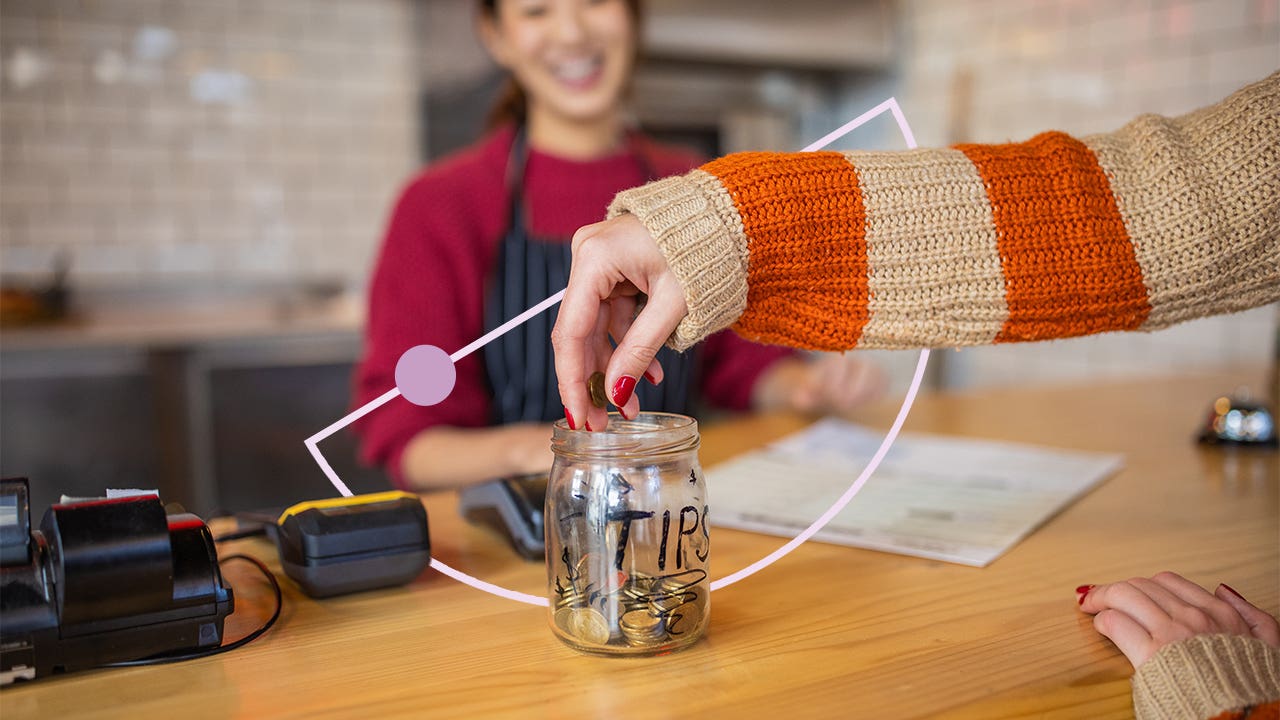 Image of a tip jar, filled with money, sitting on a counter. There is a hand adding more money to it while a smiling worker in an apron looks on.
