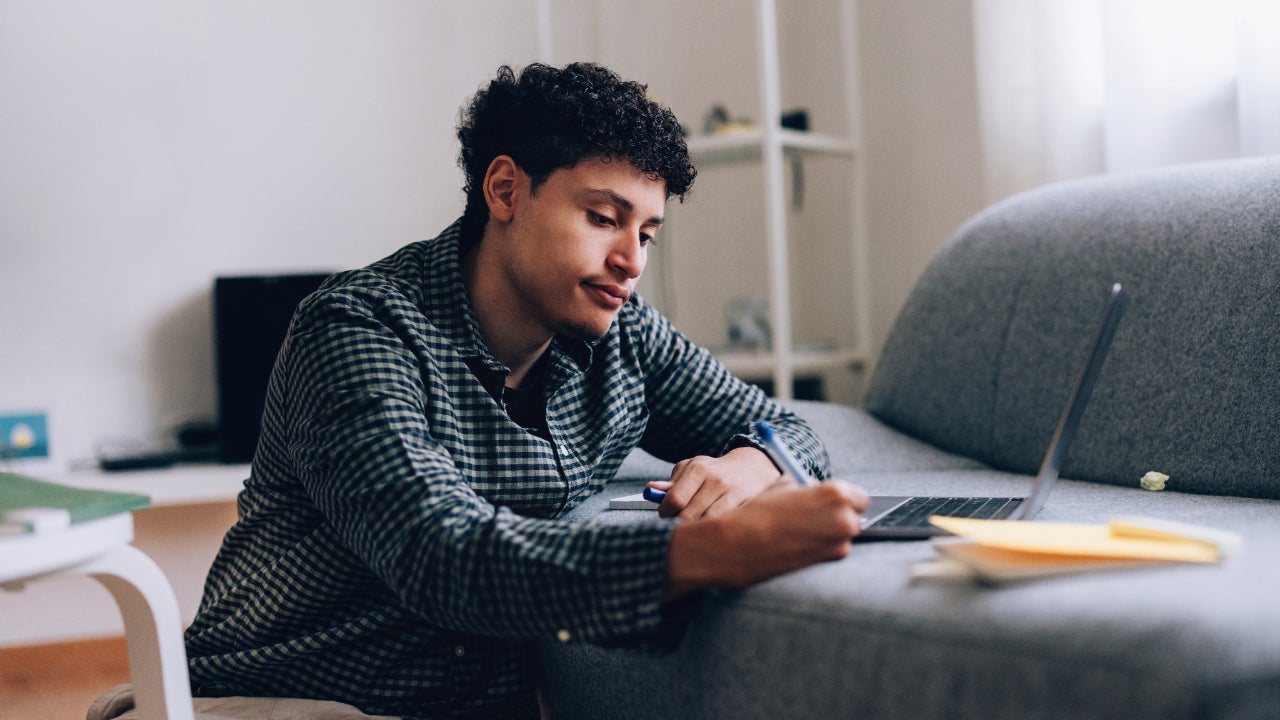 A young Black man takes notes while looking thoughtfully at his laptop.