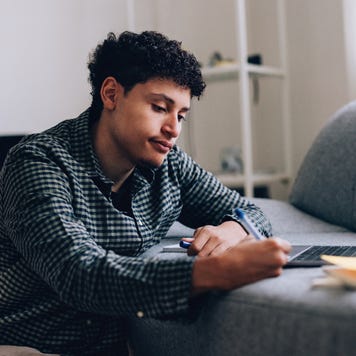 A young Black man takes notes while looking thoughtfully at his laptop.