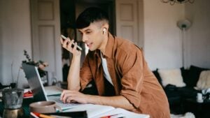Young male freelancer talking on speaker phone while working on laptop at home office